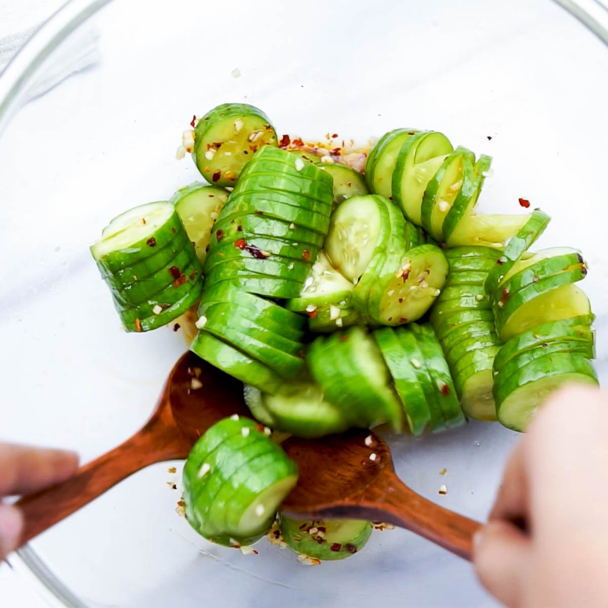 Tossing cucumber slices with spicy dressing using wooden salad spoons in a glass bowl.