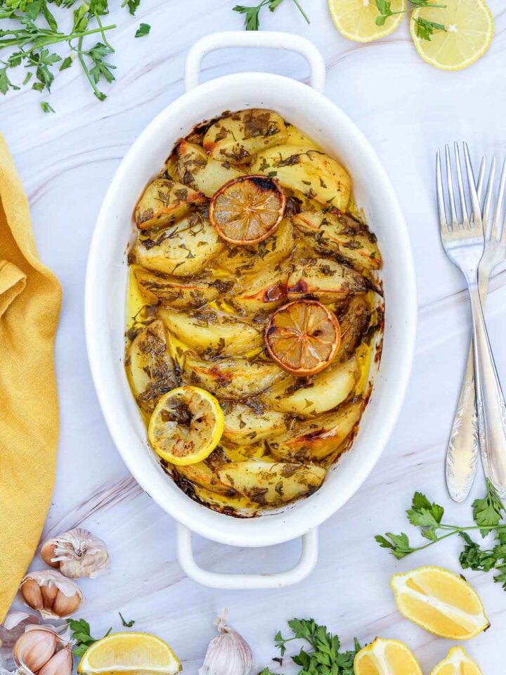 Top-down view of Greek lemon potatoes in a white oval baking dish, garnished with lemon slices and fresh parsley.