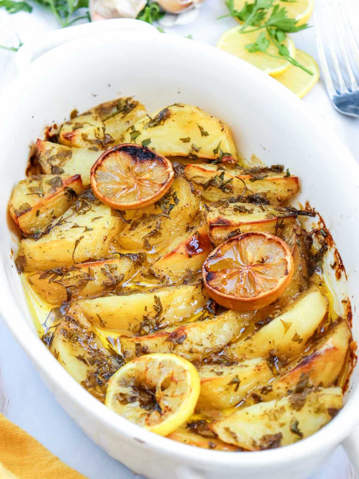 Side-angled shot of baked Greek potatoes with garlic, parsley, and lemon slices in a white casserole dish.