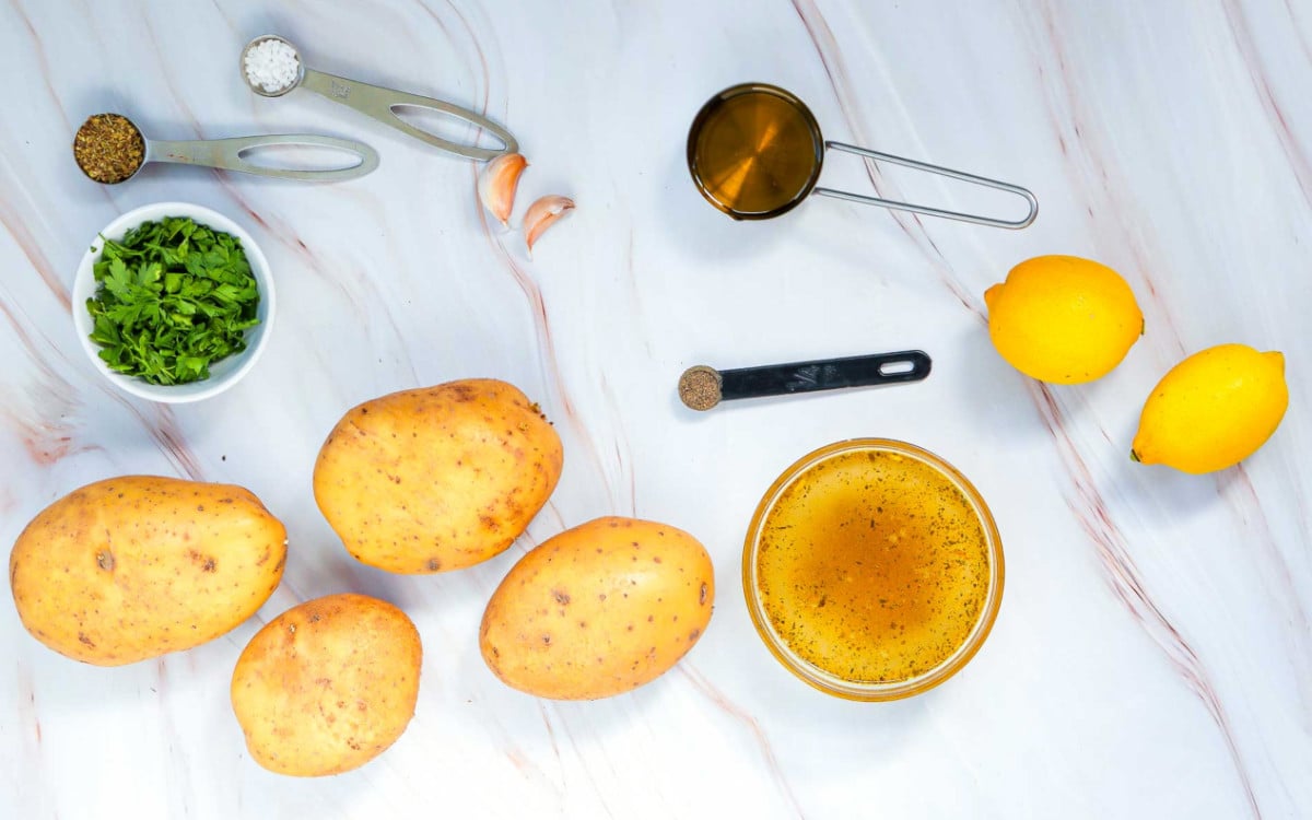Ingredients for lemon Greek potatoes arranged on a marble surface, including potatoes, lemons, garlic, parsley, olive oil, vegetable broth, and spices.