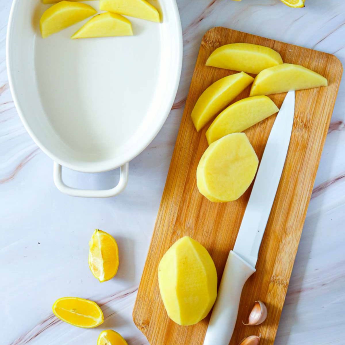 Peeled potatoes being sliced into wedges on a wooden cutting board next to a white baking dish and lemon wedges.