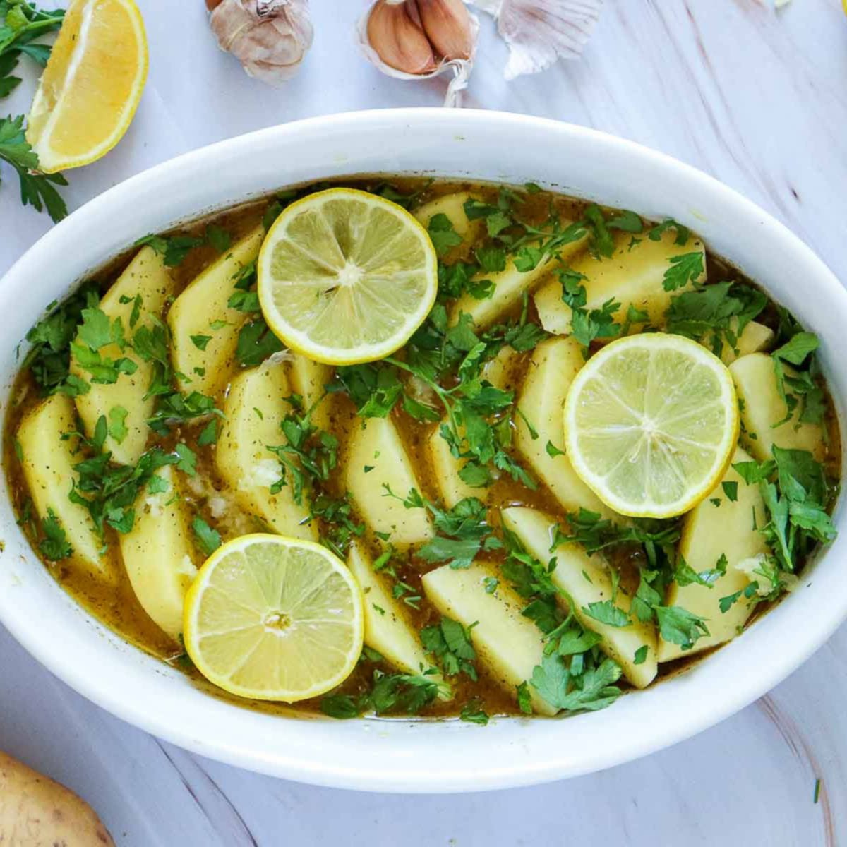 Potato wedges in marinade topped with chopped parsley, garlic, and lemon slices before baking.