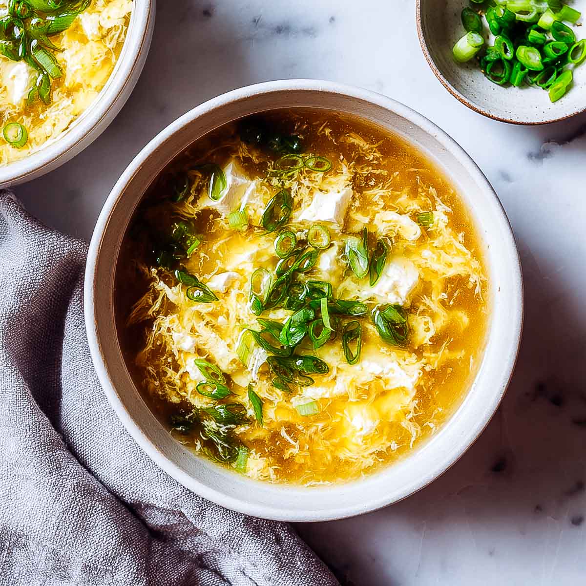 Egg drop soup with tofu cubes and scallions in a white bowl.
