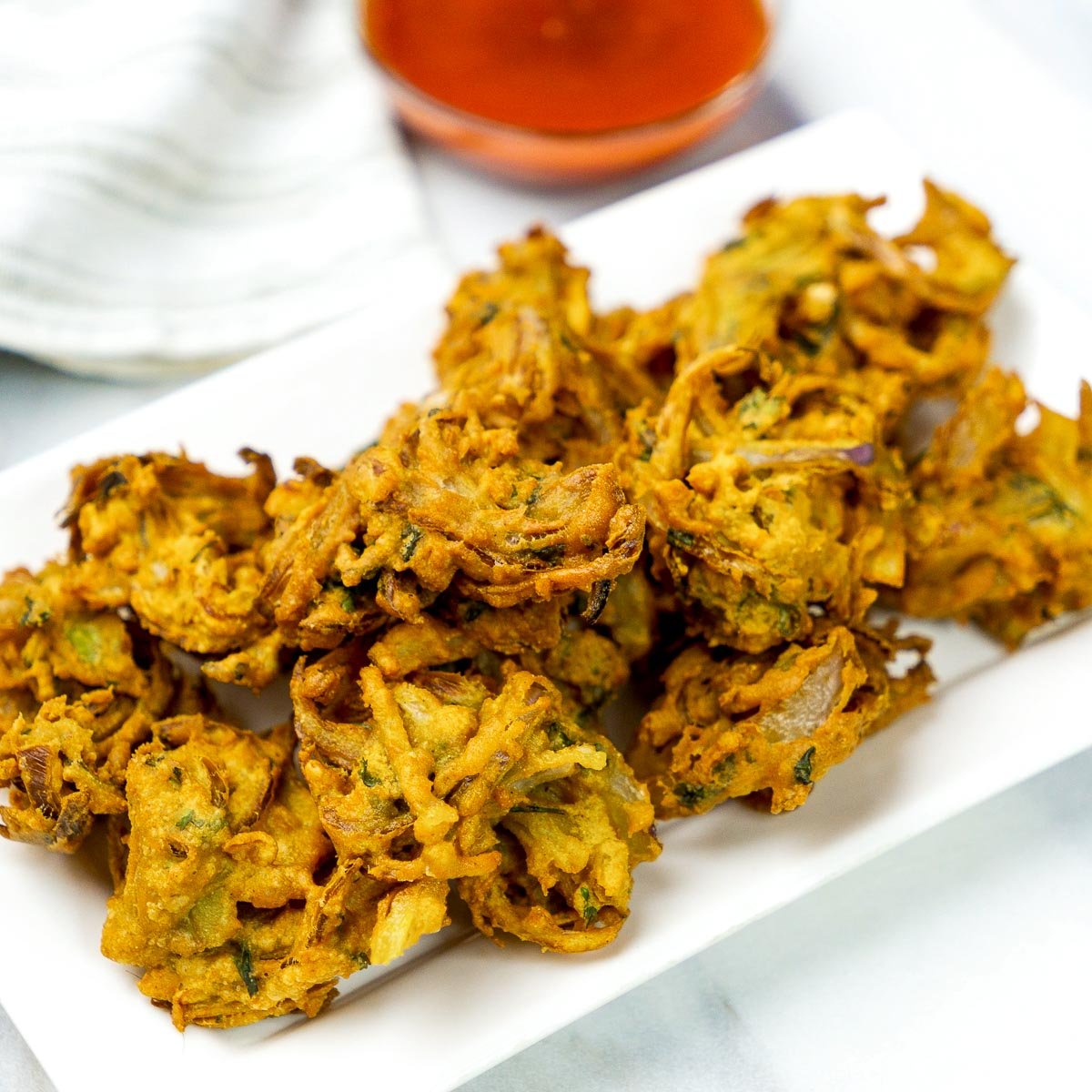 Crispy onion pakoras served on a white rectangular plate with a bowl of red chutney in the background.