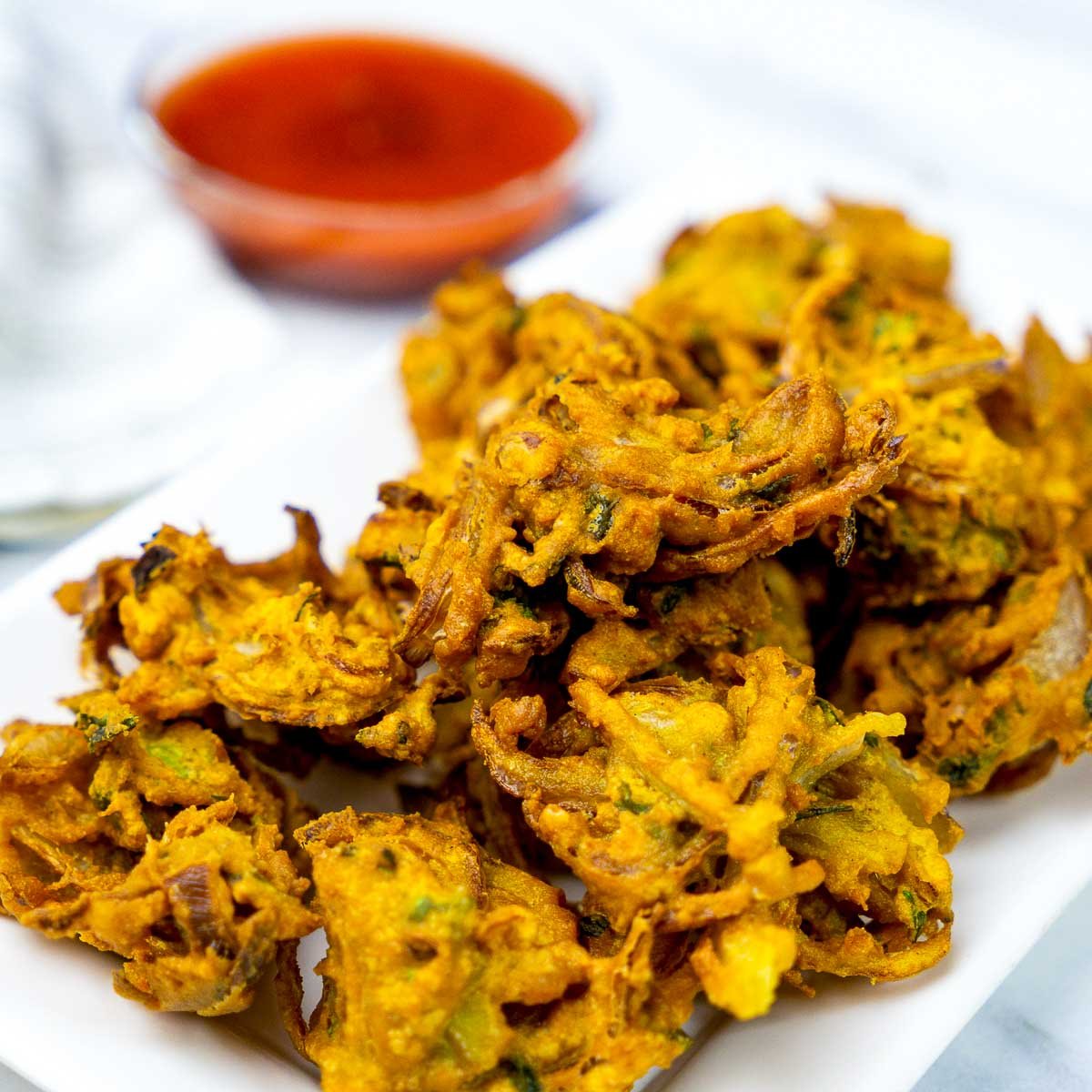 Golden brown onion fritters piled on a white plate, served with red dipping sauce.
