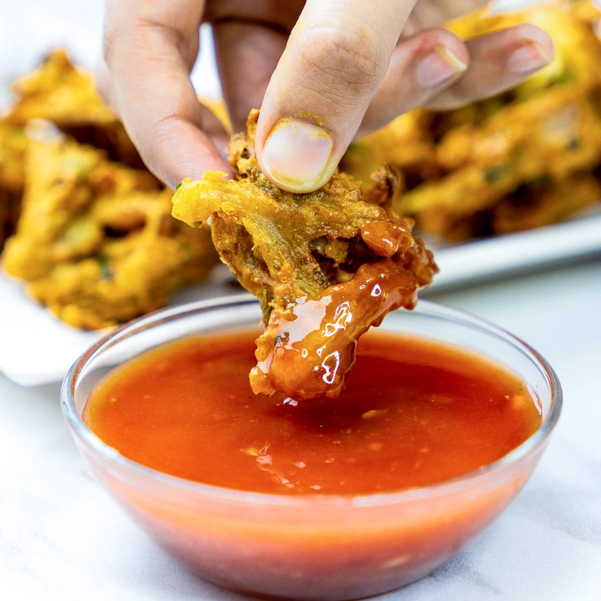 Close-up of a hand dipping a crispy onion pakora into a bowl of red chutney.