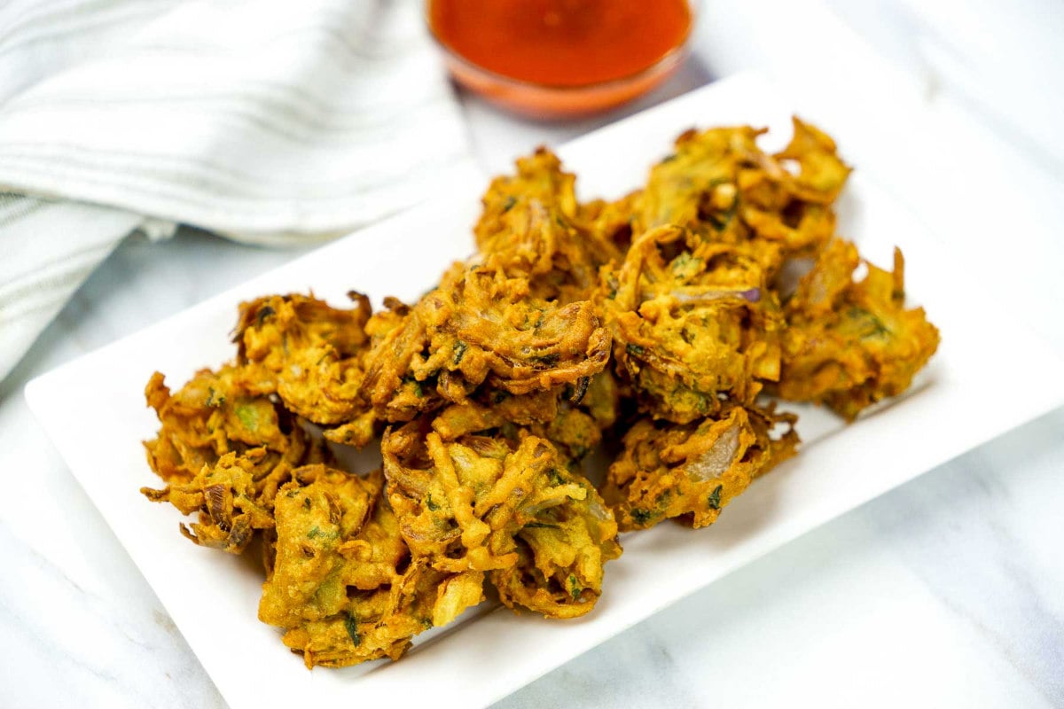 Overhead view of onion pakoras on a white serving plate with chutney and napkin in the background.