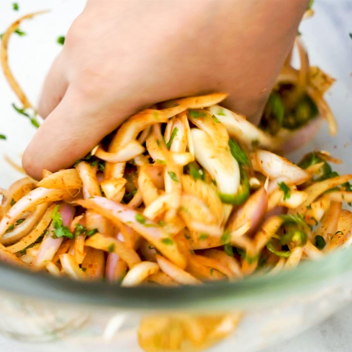 Hand mixing sliced onions, green chilies, and spices in a glass bowl for onion pakora preparation.