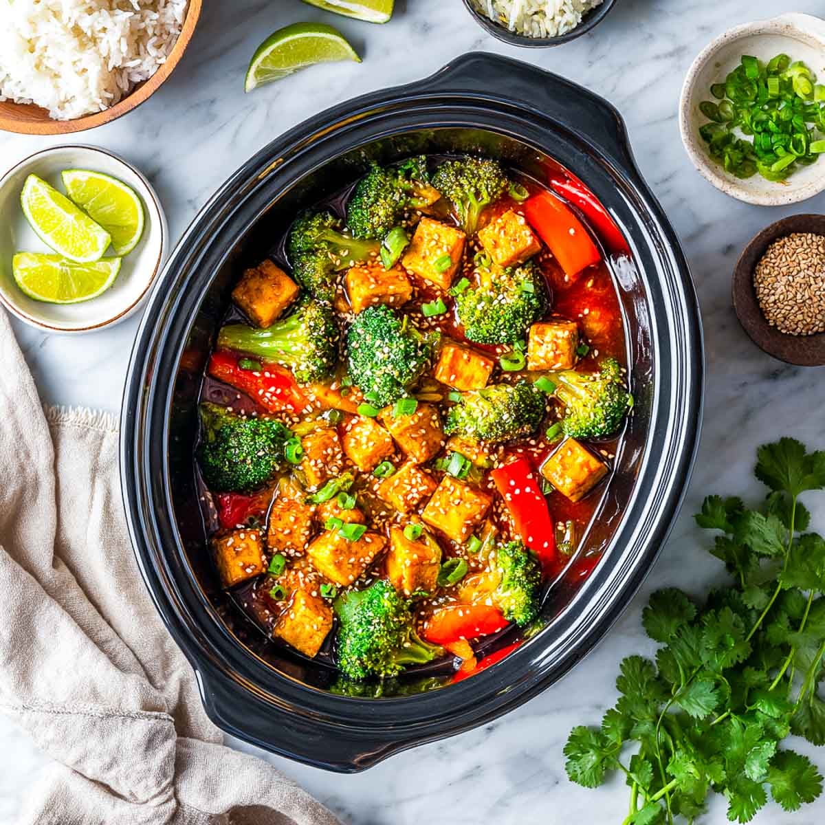 Top-down view of tofu, broccoli, red peppers, and carrots in glossy sauce inside a black slow cooker.