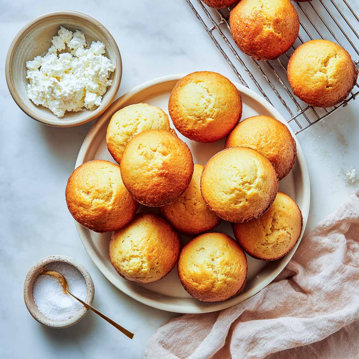 Top-down view of plain golden muffins with cottage cheese and sugar props on marble surface.