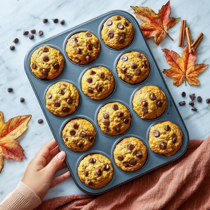 Pumpkin chocolate chip muffins in a muffin pan with fall leaves and cinnamon sticks on marble background.