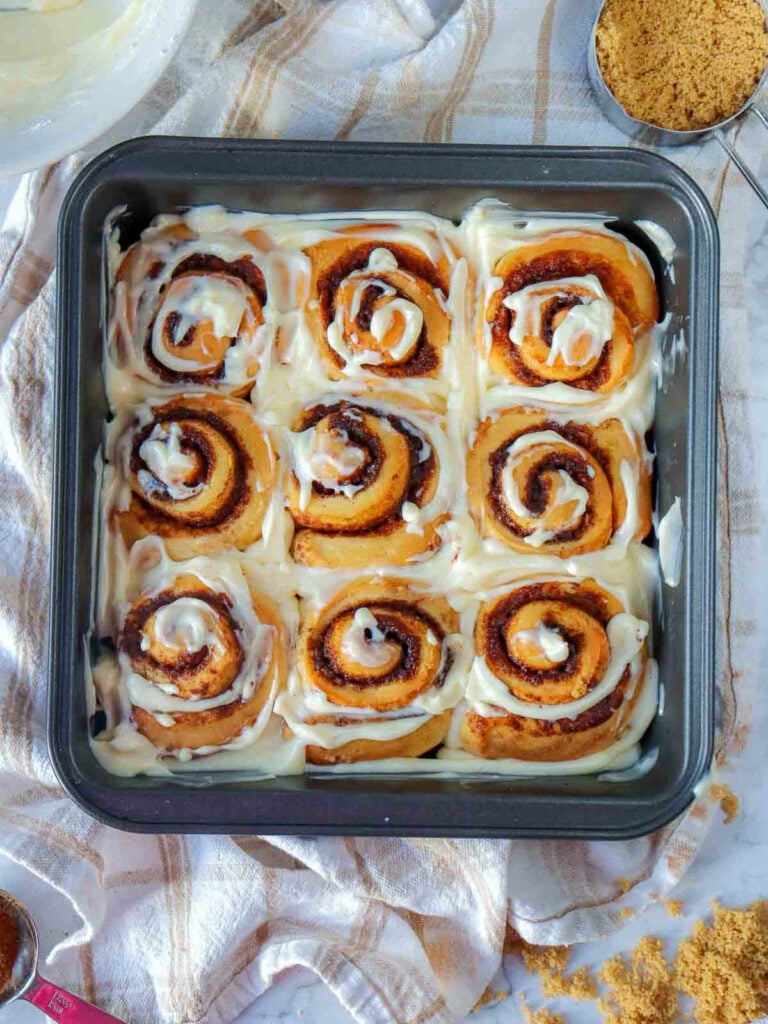 Pan of baked pumpkin cinnamon rolls covered in maple cream cheese glaze, with ingredients and linen napkin.