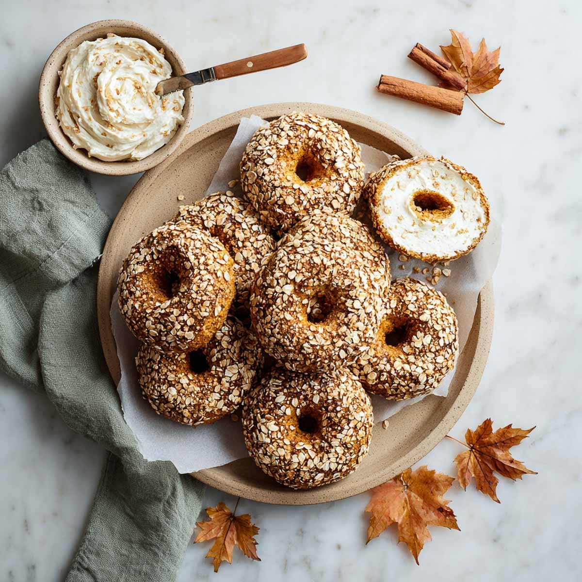 Overhead view of pumpkin spice bagels coated in oats on a rustic plate, with a bowl of maple cream cheese spread and autumn leaves on a marble background.
