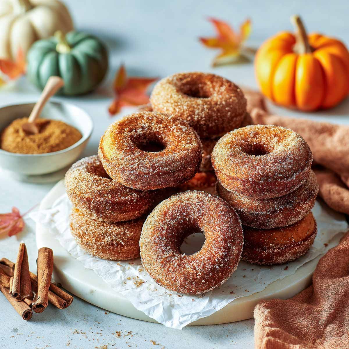 Stacked pumpkin spice donuts coated in cinnamon sugar on a parchment-lined marble board with mini pumpkins in the background.