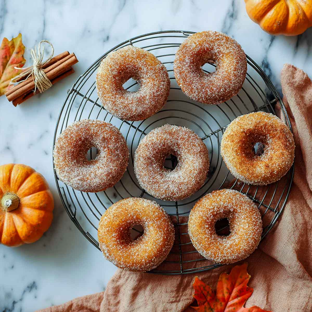 Pumpkin spice donuts arranged on a round wire cooling rack with mini pumpkins and fall leaves on marble.