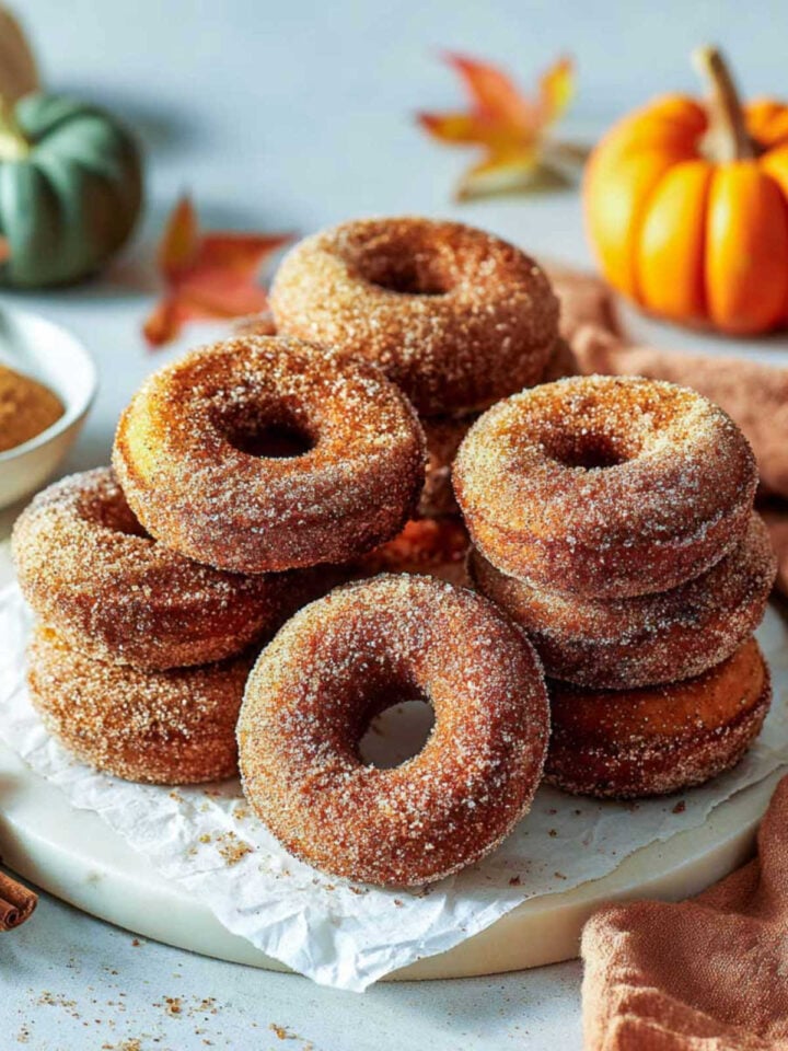 Close-up of stacked pumpkin spice donuts covered in cinnamon sugar on a parchment-lined board with autumn decor.