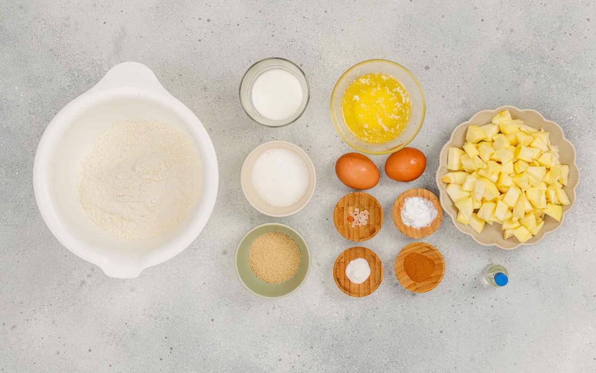 Apple cinnamon crumb muffin ingredients including flour, butter, eggs, milk, sugar, and chopped apples arranged on a counter.