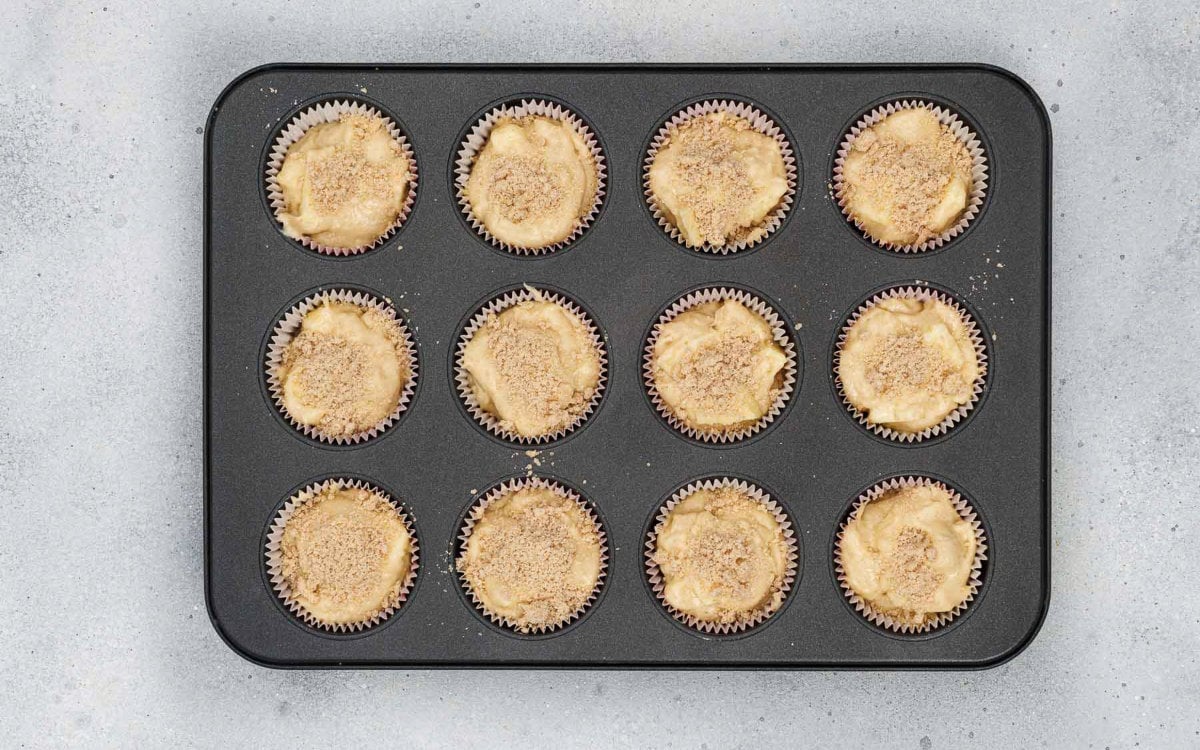 Muffin tin filled with apple cinnamon muffin batter topped with crumb mixture before baking.
