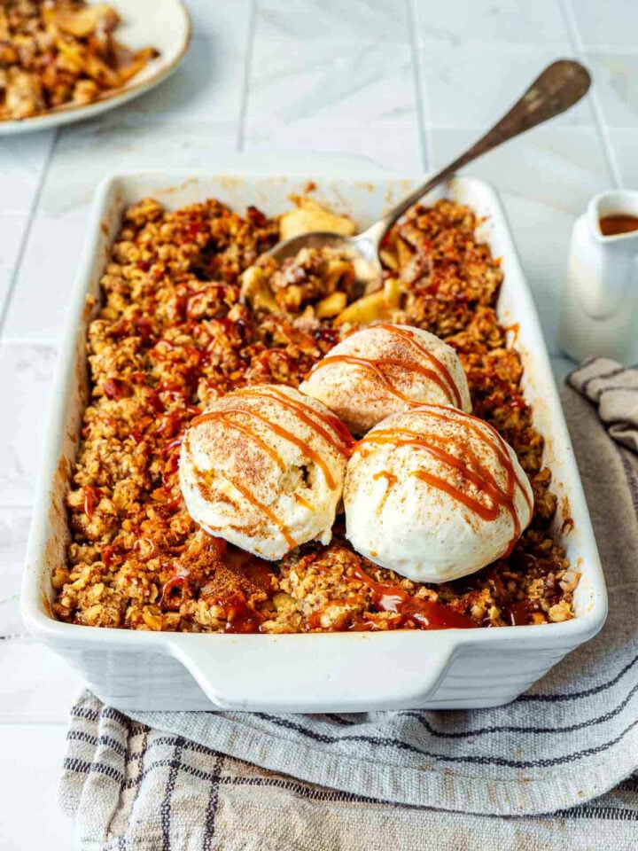 Apple crisp in a white baking dish topped with vanilla ice cream scoops and caramel drizzle.