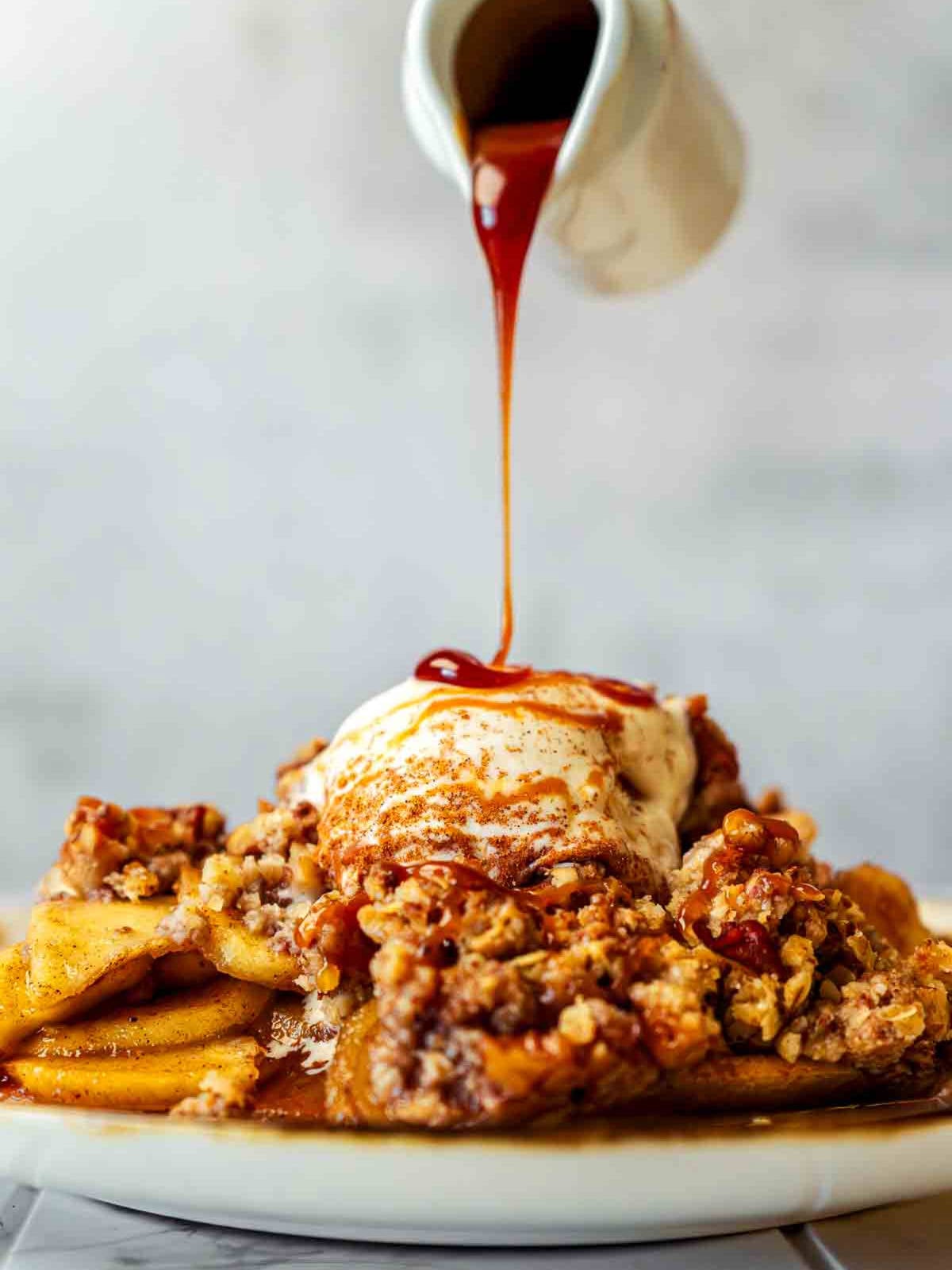 Caramel sauce being poured over warm apple crisp topped with vanilla ice cream.