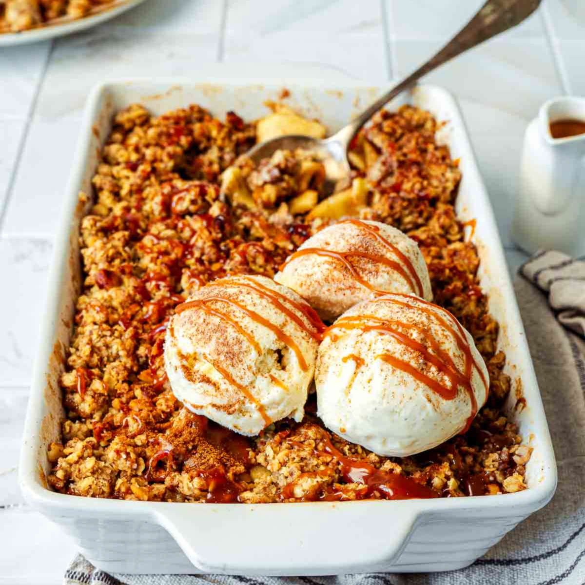 Apple crisp in a white baking dish topped with vanilla ice cream scoops and caramel drizzle.