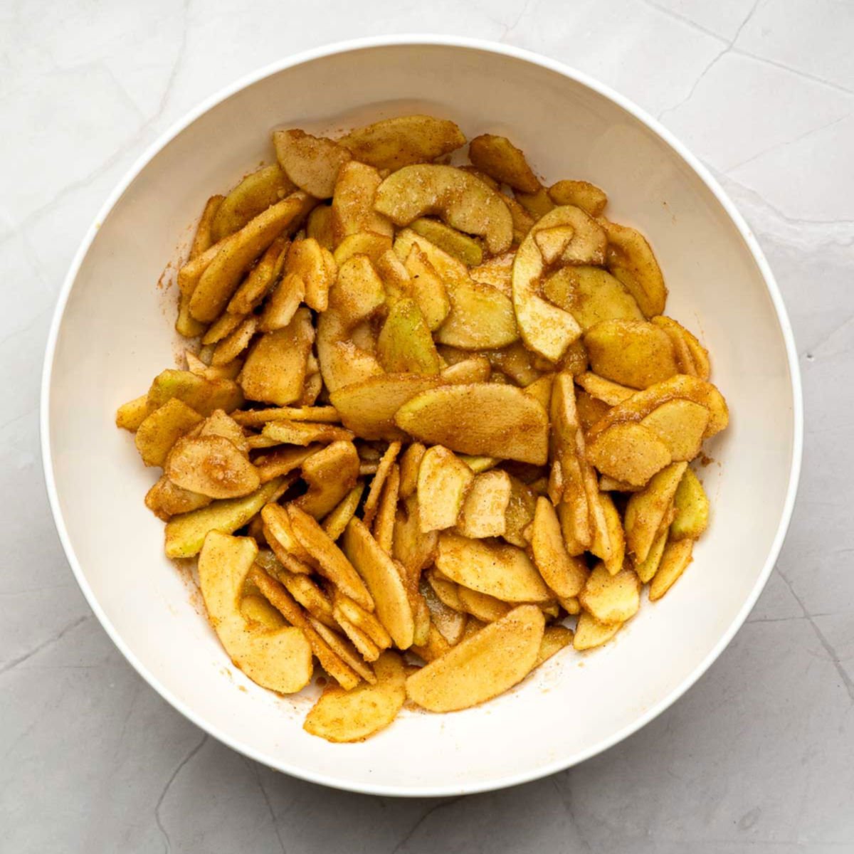 Sliced apples tossed with cinnamon, sugar, and spices in a mixing bowl.