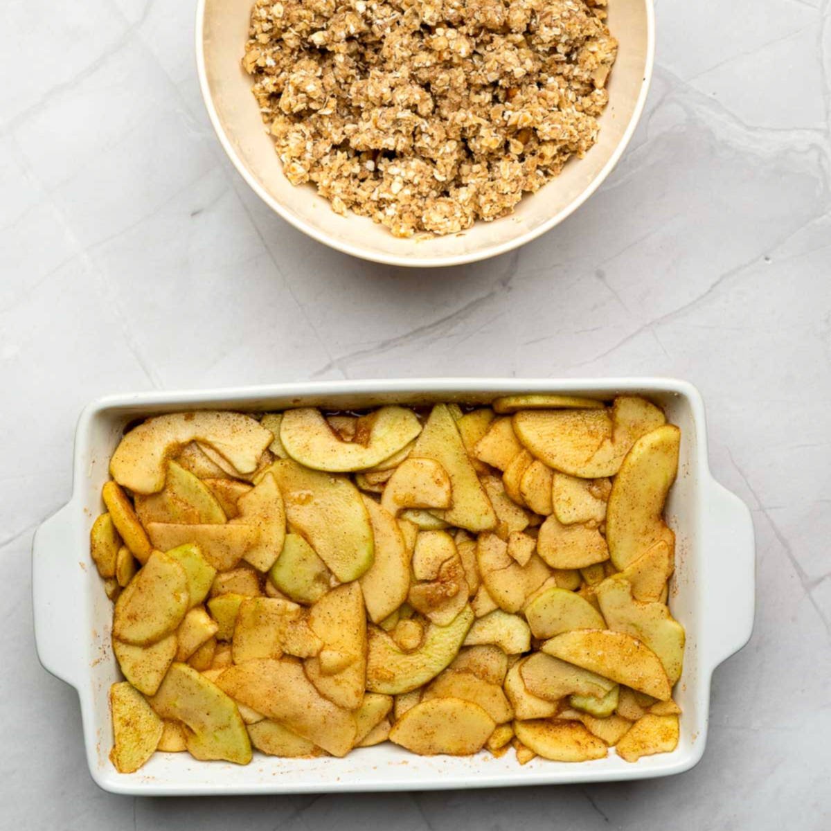 Baking dish filled with spiced apple slices with crisp topping mixture in a bowl nearby.