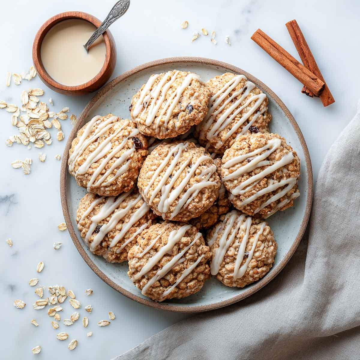 Plate of apple oatmeal raisin cookies drizzled with cinnamon glaze, surrounded by oats and cinnamon sticks.