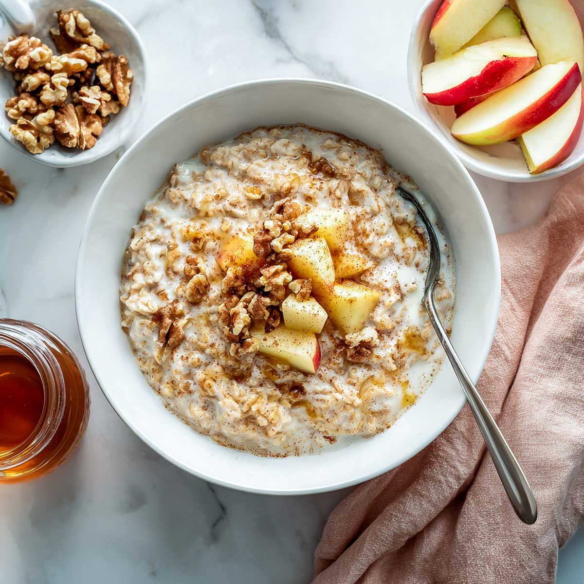 A white bowl of creamy apple cinnamon oatmeal topped with fresh apple slices, walnuts, and cinnamon, with a jar of maple syrup on the side.