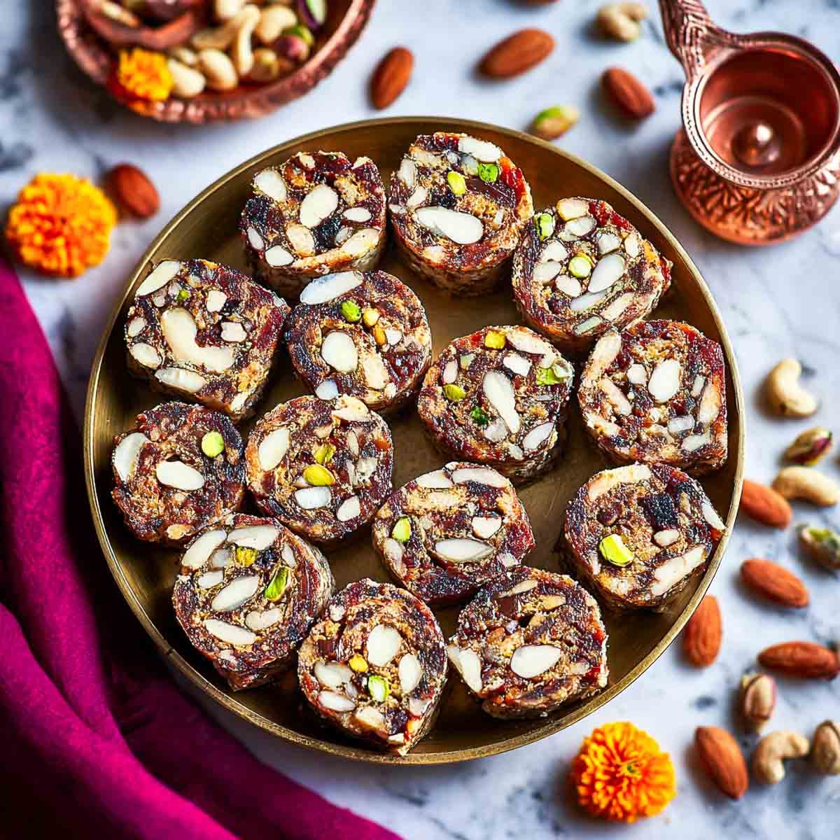 Round brass plate filled with sliced date and nut rolls, surrounded by assorted nuts.