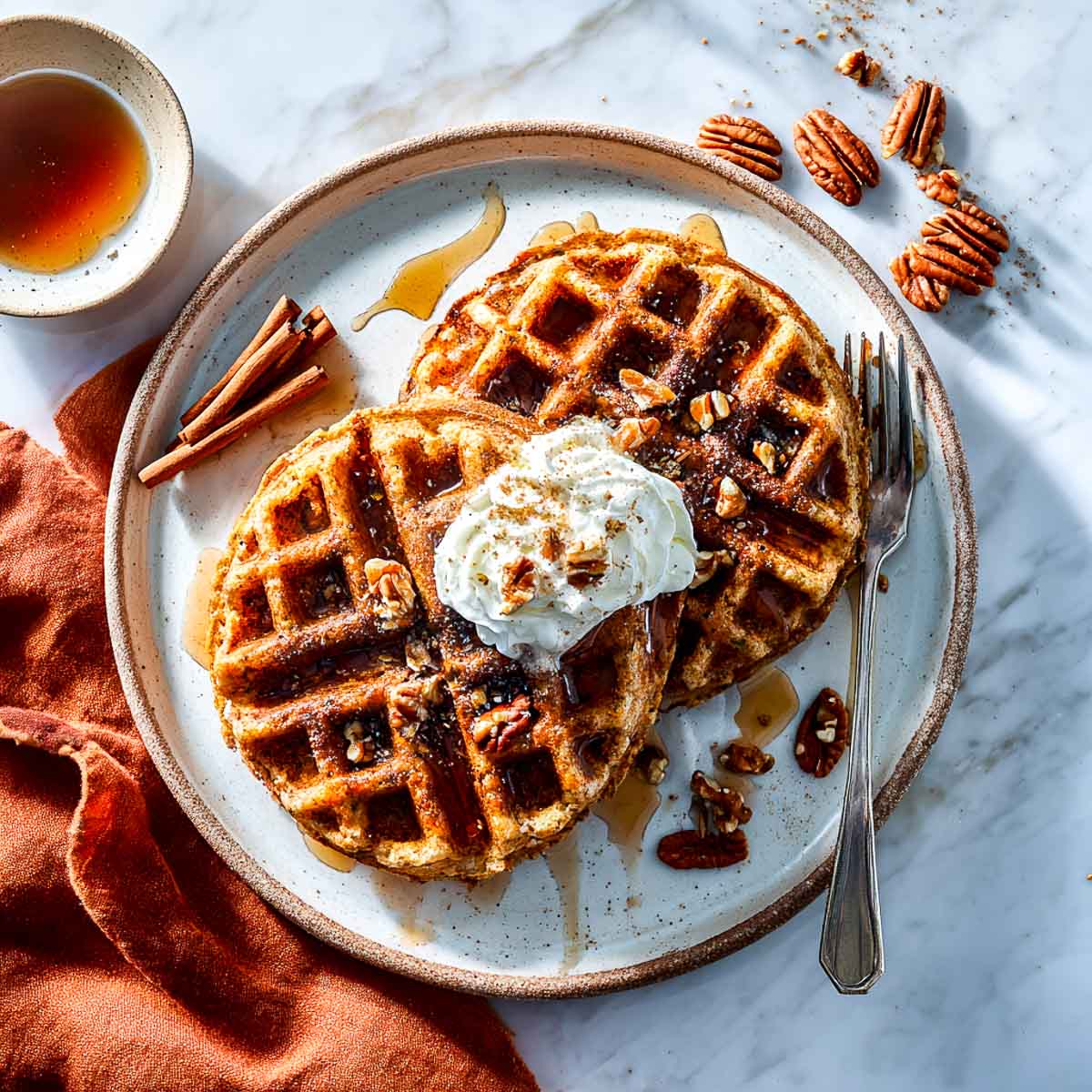 Two golden-brown cottage cheese waffles on a ceramic plate topped with whipped cream, chopped pecans, and maple syrup, with cinnamon sticks on the side.