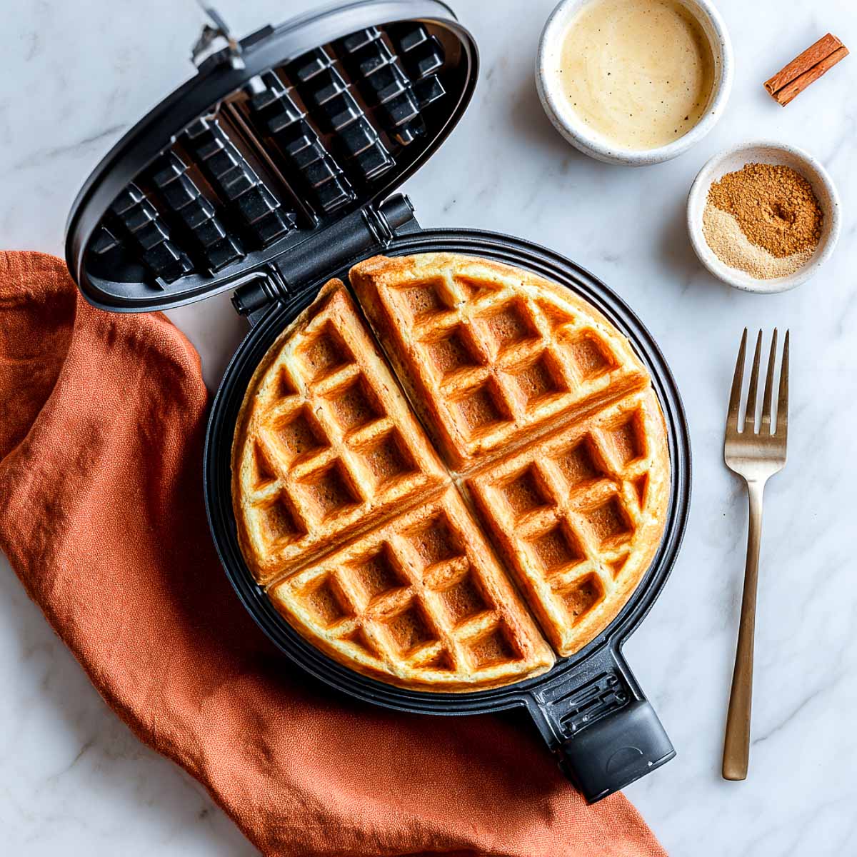 Freshly cooked cottage cheese waffle in a waffle maker, with cinnamon and sugar in small bowls nearby.