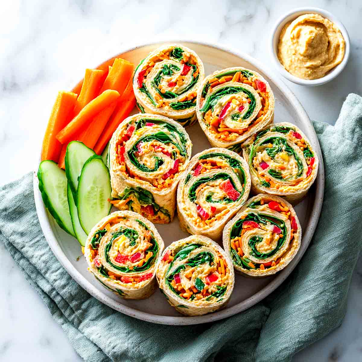 Veggie hummus pinwheels on a plate with cucumber, carrots, and dip.