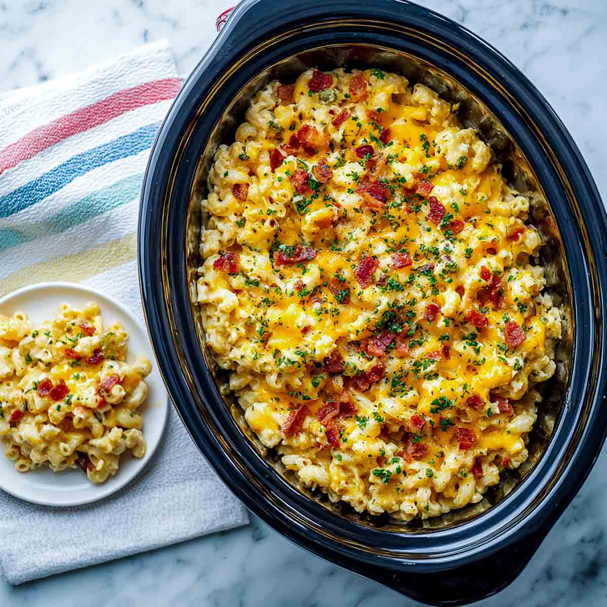 Top-down view of creamy breakfast mac and cheese in a black oval crockpot, served with toast and herbs on a white marble background.