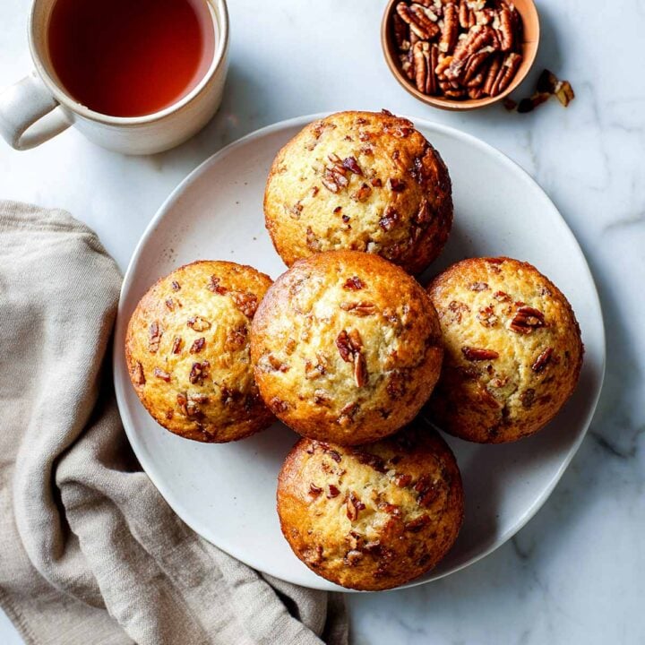 Overhead view of five maple pecan cottage cheese muffins on a white plate, served with a cup of tea and a small bowl of pecans.