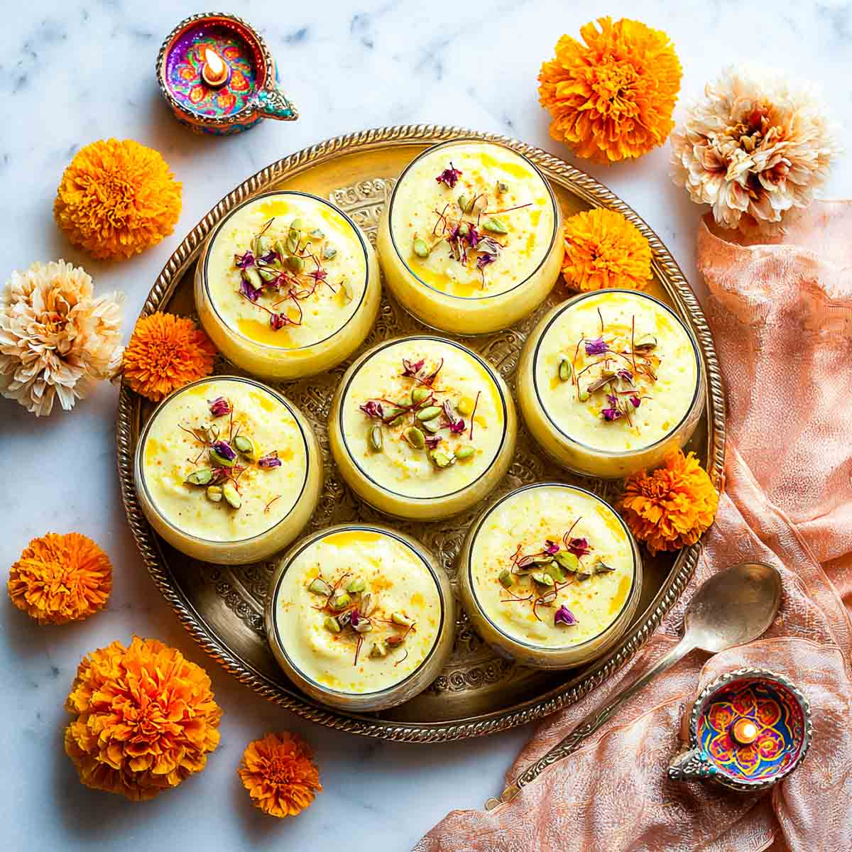 Overhead view of Ras Malai mousse cups with pistachios, saffron, and rose petals on a brass tray.
