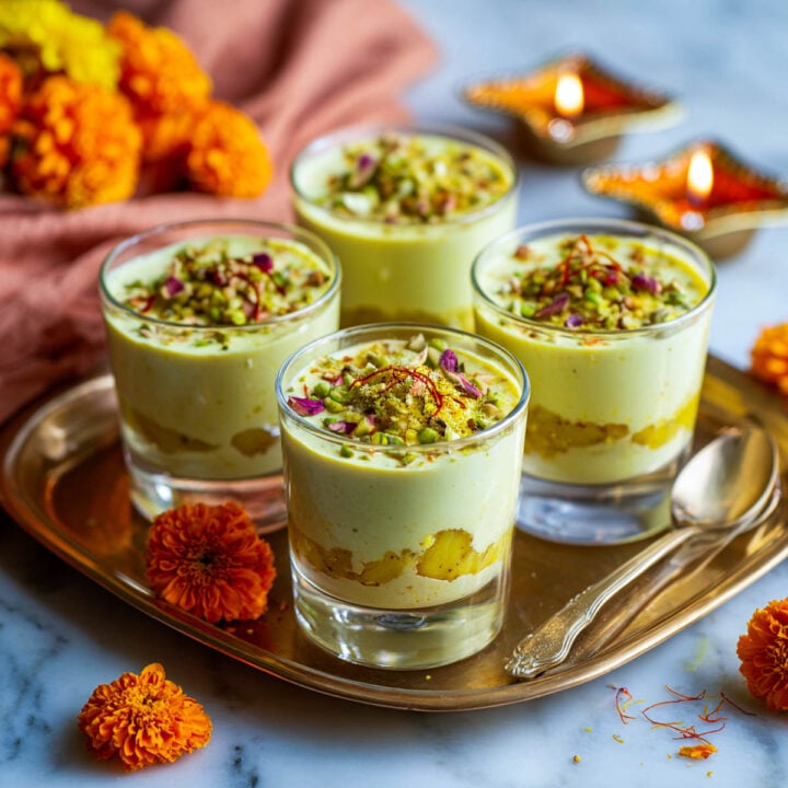 Four glass cups of Ras Malai mousse with pistachios, saffron, and rose petals on a brass tray.