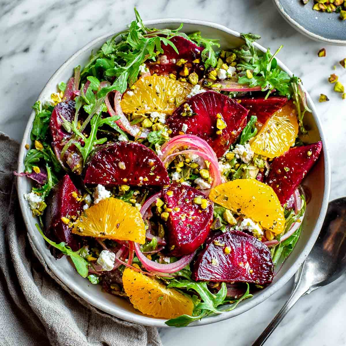 Roasted beet and citrus salad with vegan feta, pistachios, and arugula in a white bowl on a marble surface.