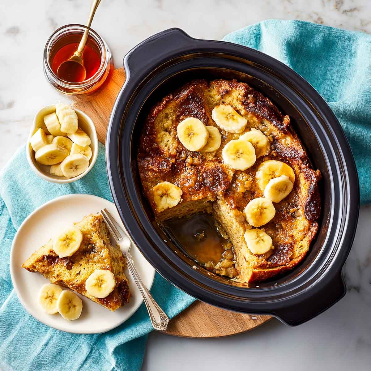 Top-down view of banana French toast casserole in a black crockpot with banana slices on top and a portion served on a white plate.