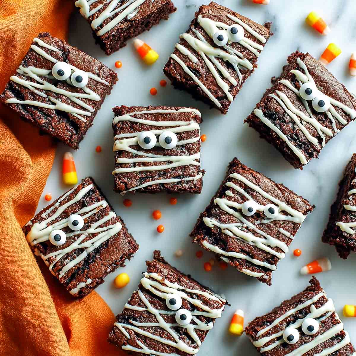 Fudgy Halloween mummy brownies drizzled with white chocolate and topped with candy eyes, surrounded by candy corn.
