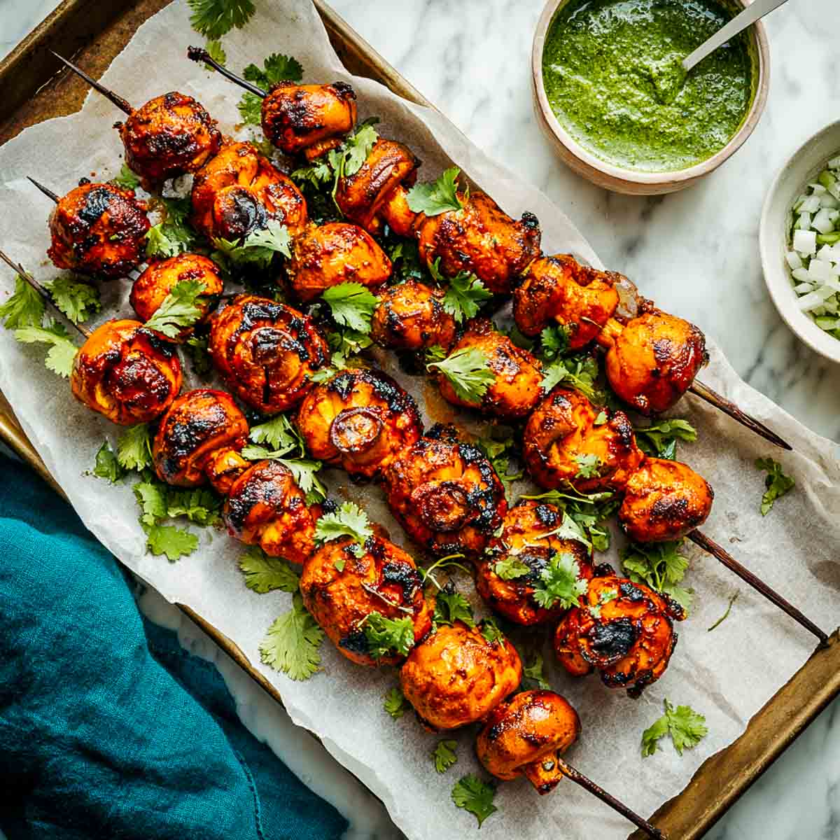 Overhead view of skewered tandoori mushrooms on a tray lined with parchment paper, garnished with cilantro, and accompanied by bowls of green chutney and diced onions.