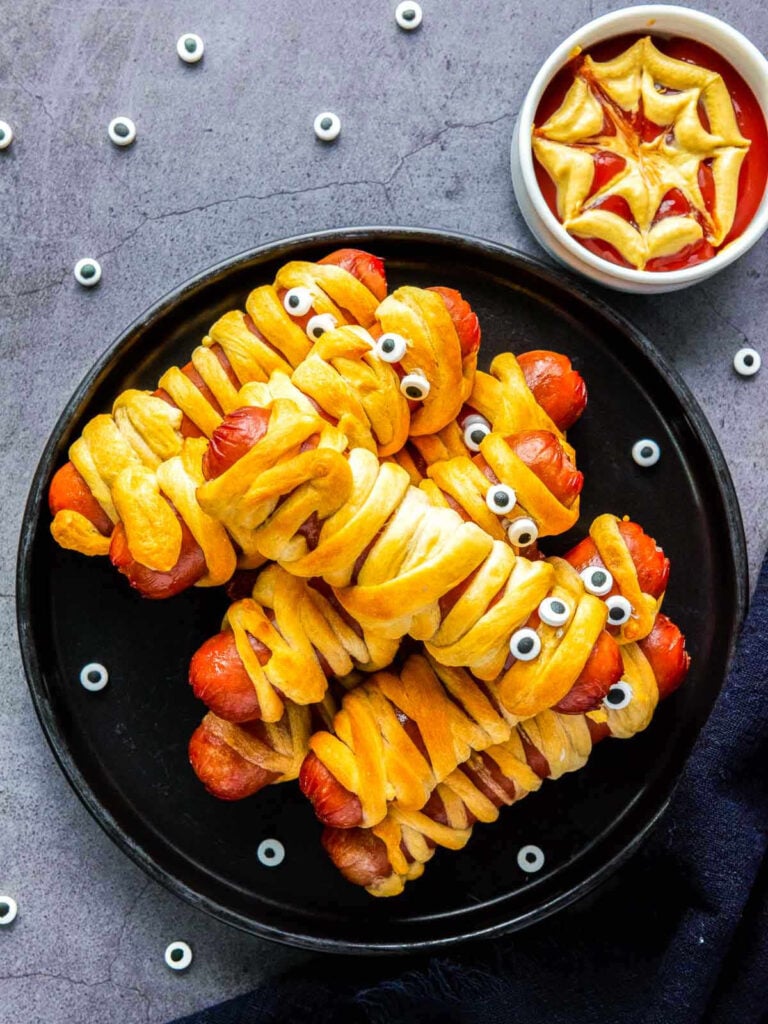 Halloween mummy hot dogs on black plate with spiderweb ketchup dip.