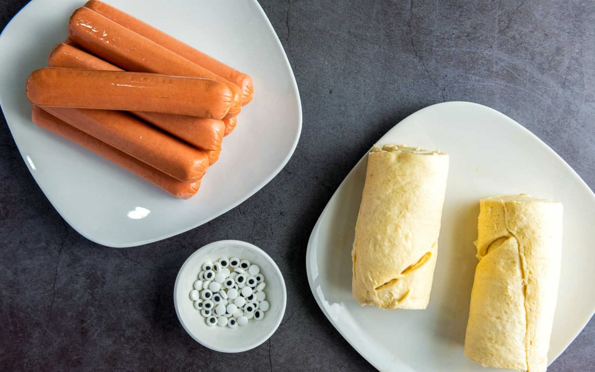 Hot dogs, crescent roll dough, and candy eyes displayed on white plates.