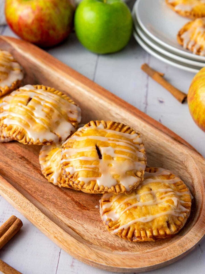 Freshly baked apple hand pies with icing drizzle displayed on a wooden tray next to apples and cinnamon sticks.