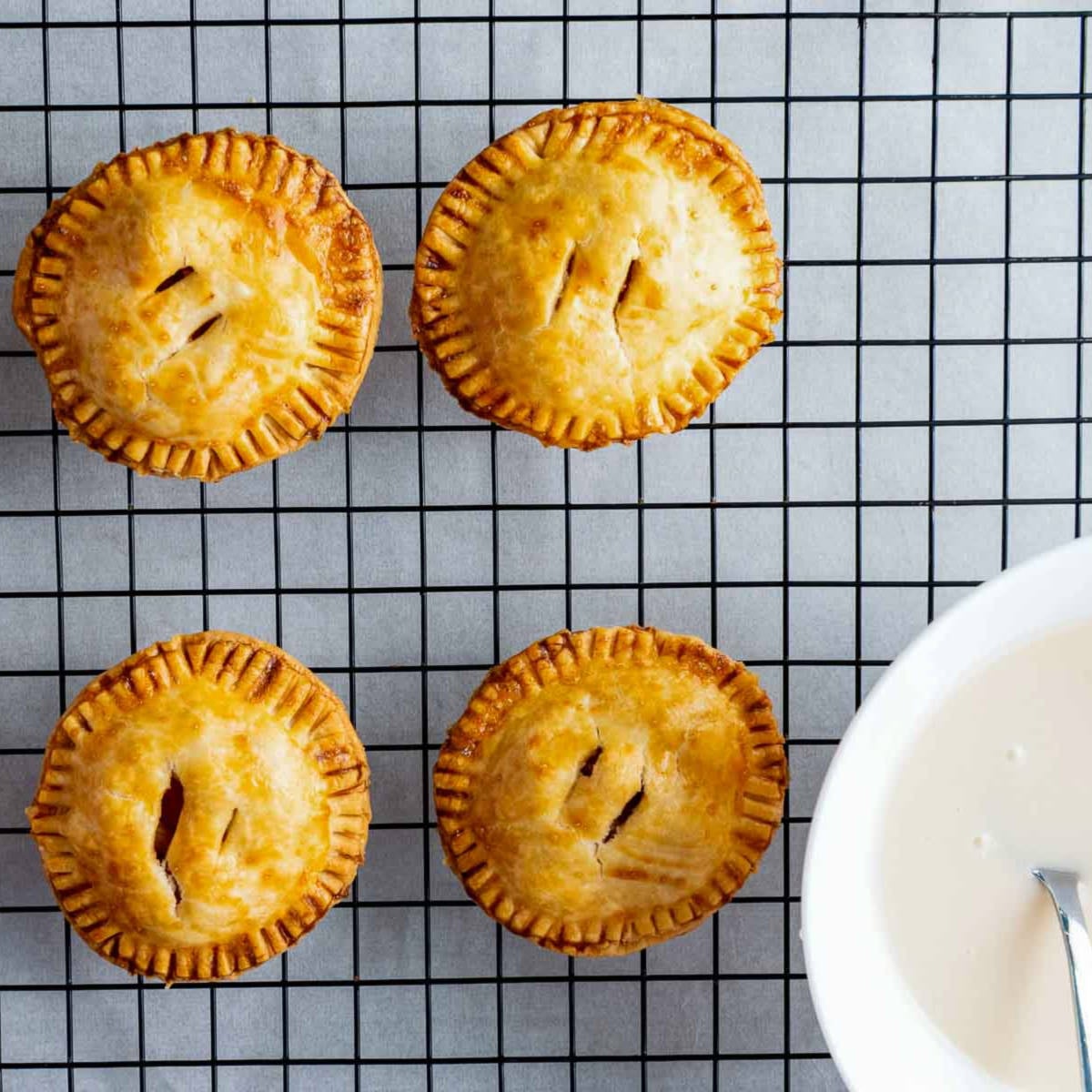 Apple hand pies cooling on a wire rack with a bowl of icing on the side.
