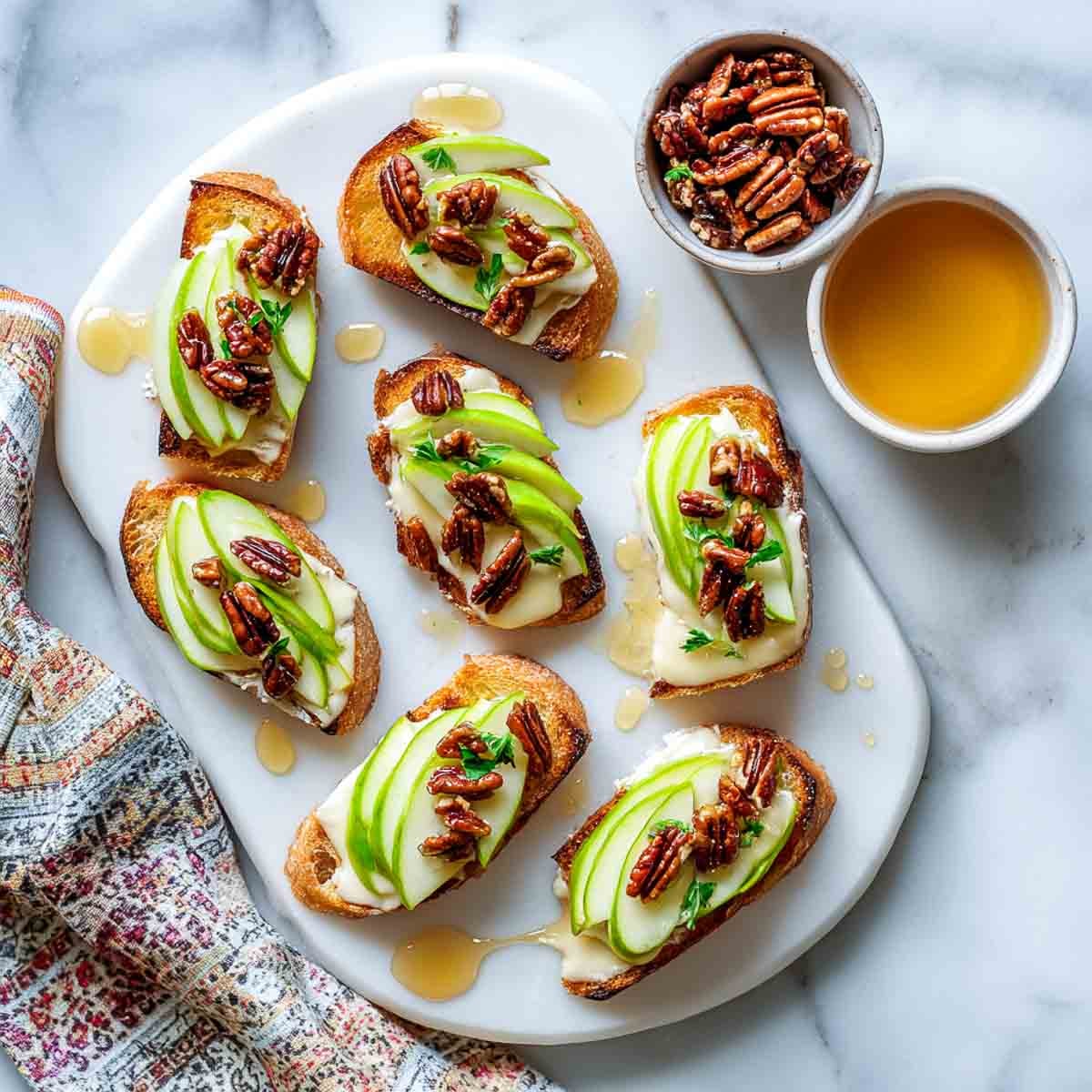 Crostini topped with melted brie, apple slices, maple pecans, and a maple drizzle on marble platter.