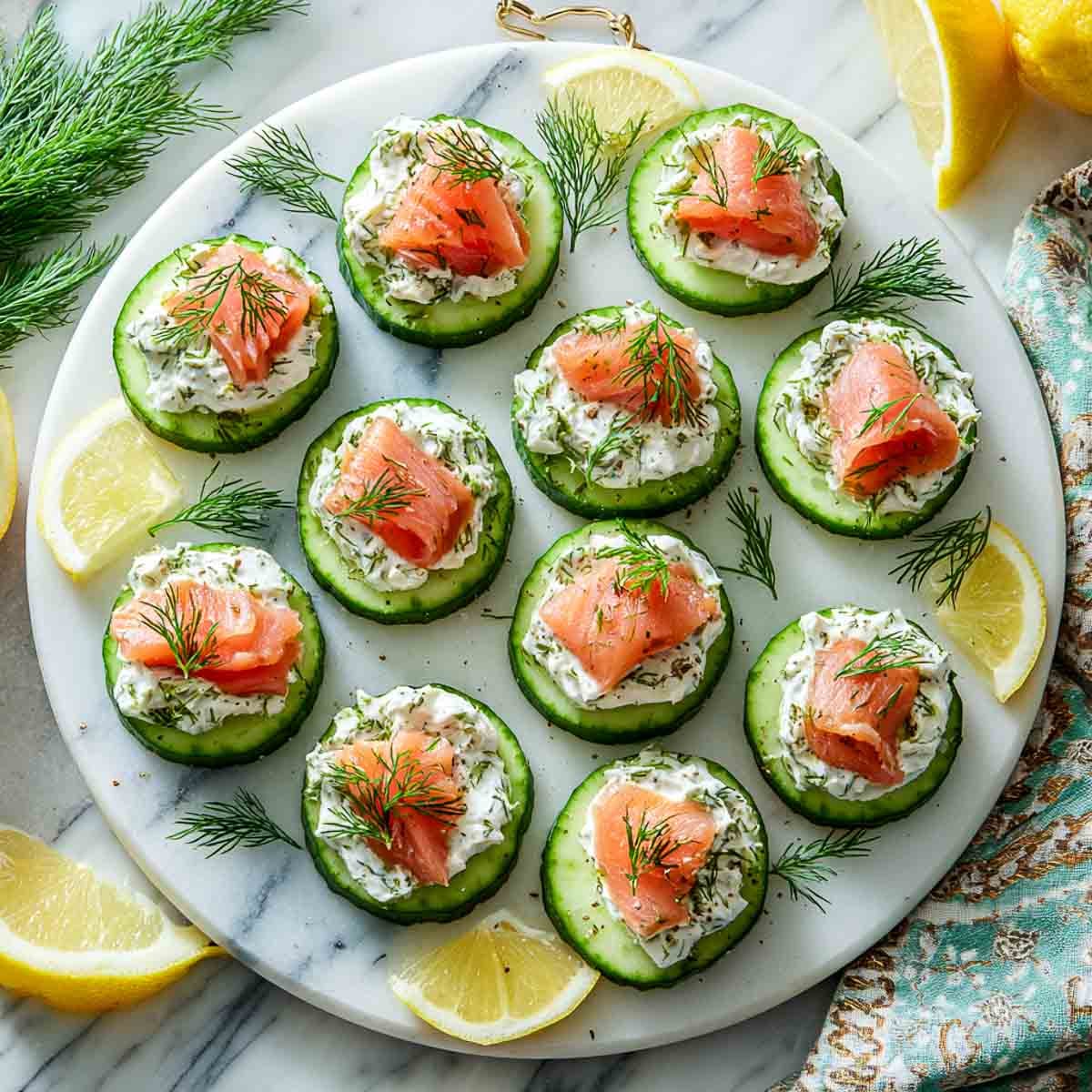 Cucumber slices topped with cream cheese, smoked salmon, and dill on a white platter with lemon wedges.