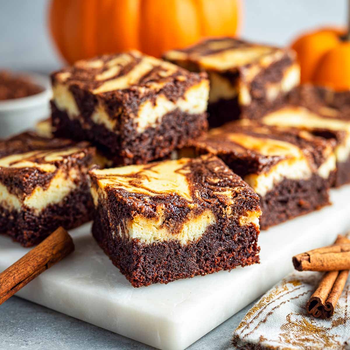 Gluten-free pumpkin cheesecake swirl brownies cut into neat squares on a marble board with cinnamon sticks.