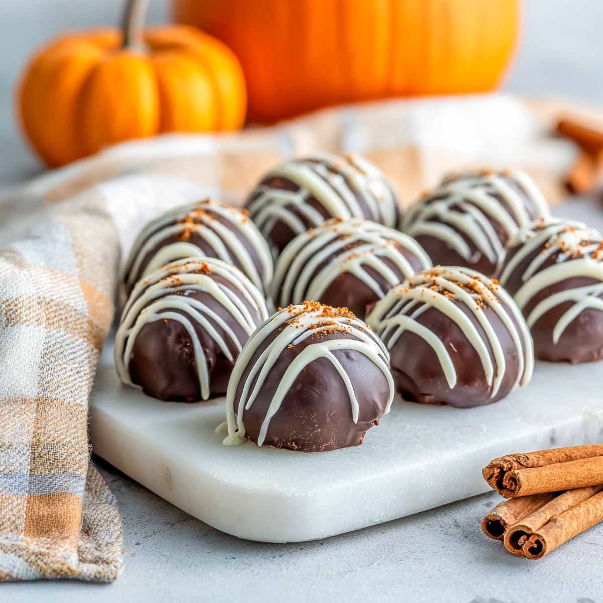 Gluten-free pumpkin spice truffles coated in chocolate with a white drizzle, arranged neatly on a marble board with cinnamon sticks and pumpkins.