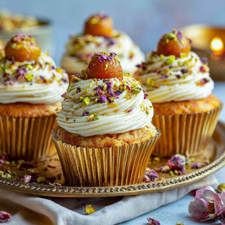Close-up of Gulab Jamun stuffed cupcakes topped with whipped frosting, crushed pistachios, rose petals, and a mini gulab jamun on top.
